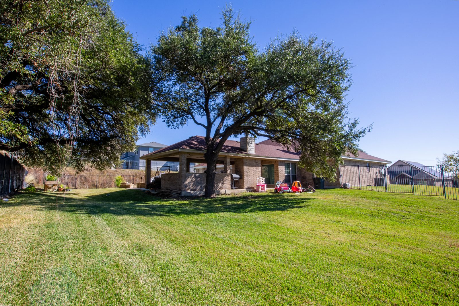 Wide green lawn shaded by large oak trees behind the brick home and covered patio, with an iron fence around the yard