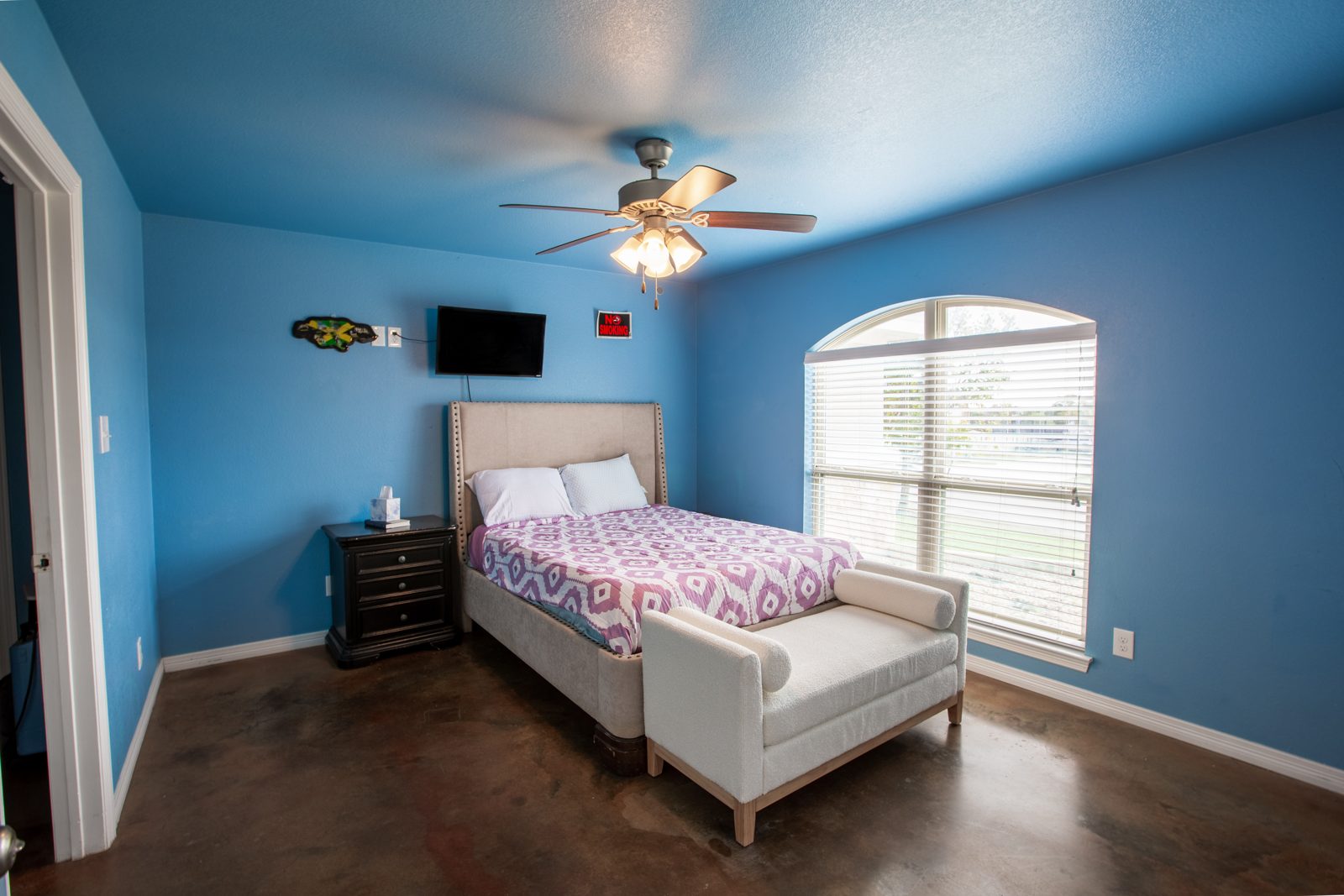 Blue guest bedroom with an upholstered queen bed, white bench at the foot, nightstand, arched window and wall TV