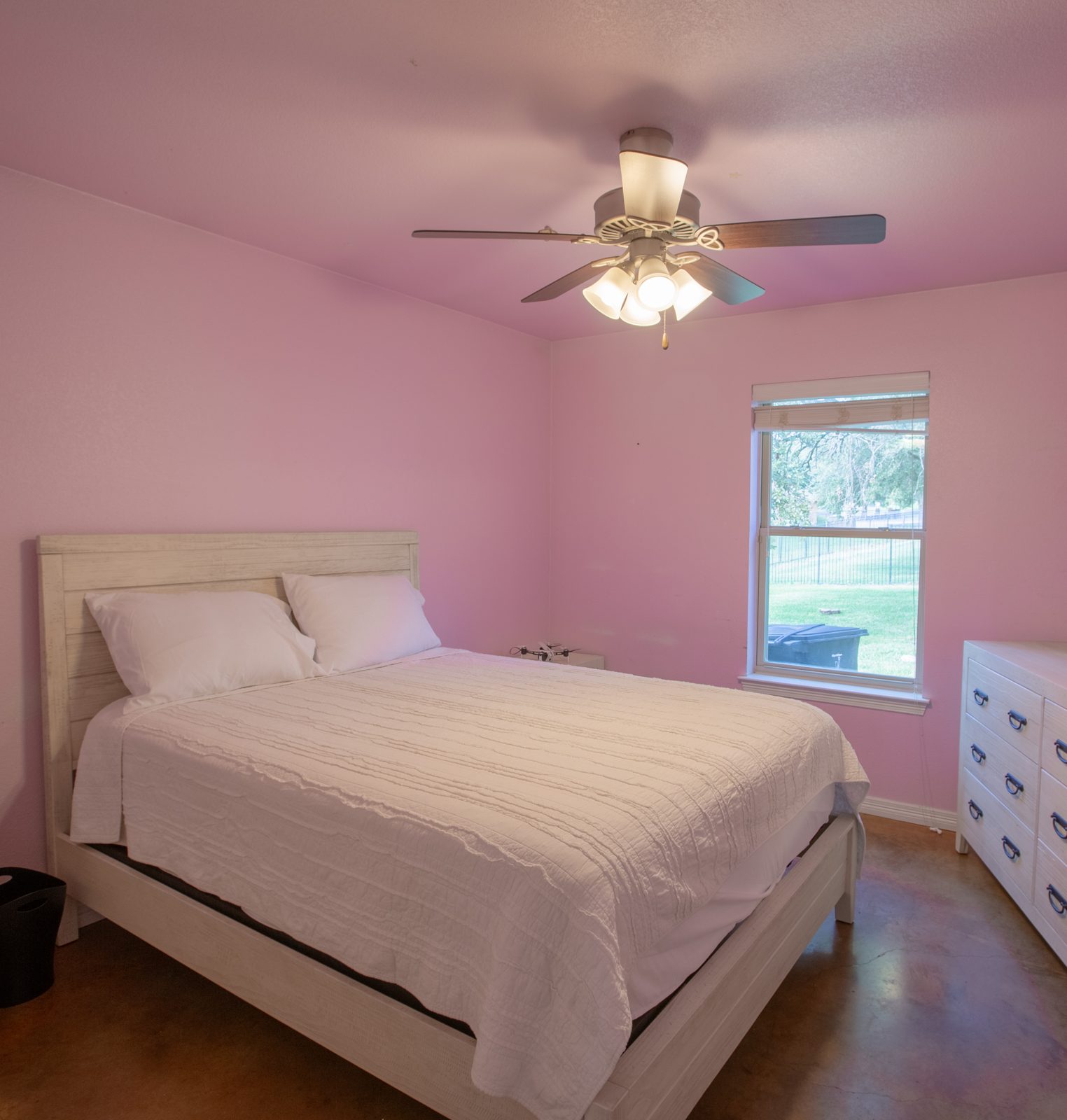 Pink guest bedroom with a queen bed in white bedding, dresser with mirror, ceiling fan and wall TV