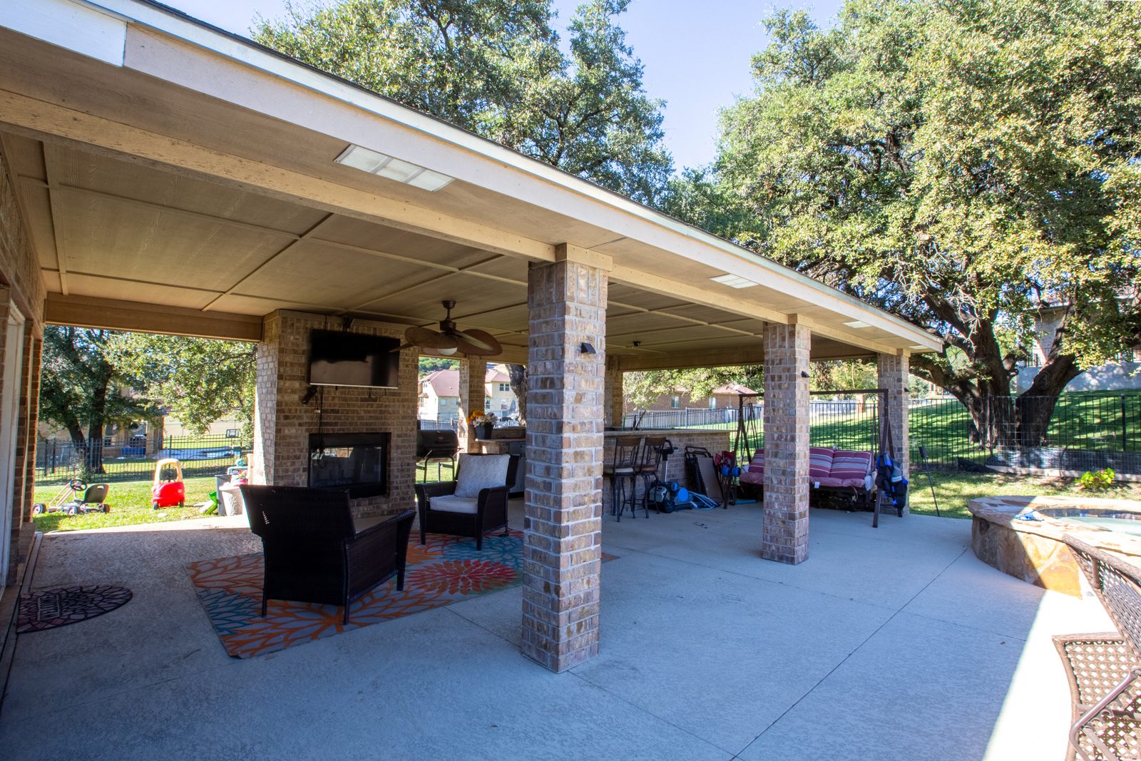 Large covered brick patio with a wood-burning fireplace and wall TV, wicker chairs on a bright rug, bar seating and a porch swing, shaded by oak trees