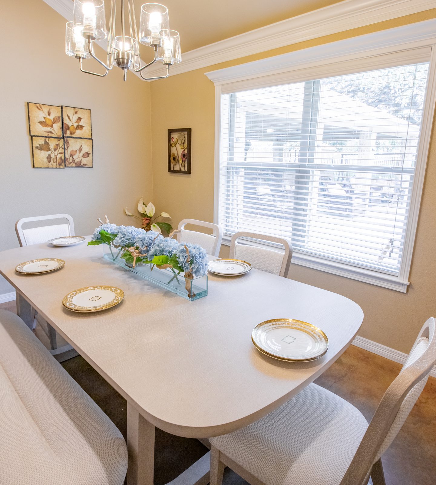 Dining table with bench and upholstered chairs, gold-rimmed plates and a blue hydrangea centerpiece under a modern chandelier