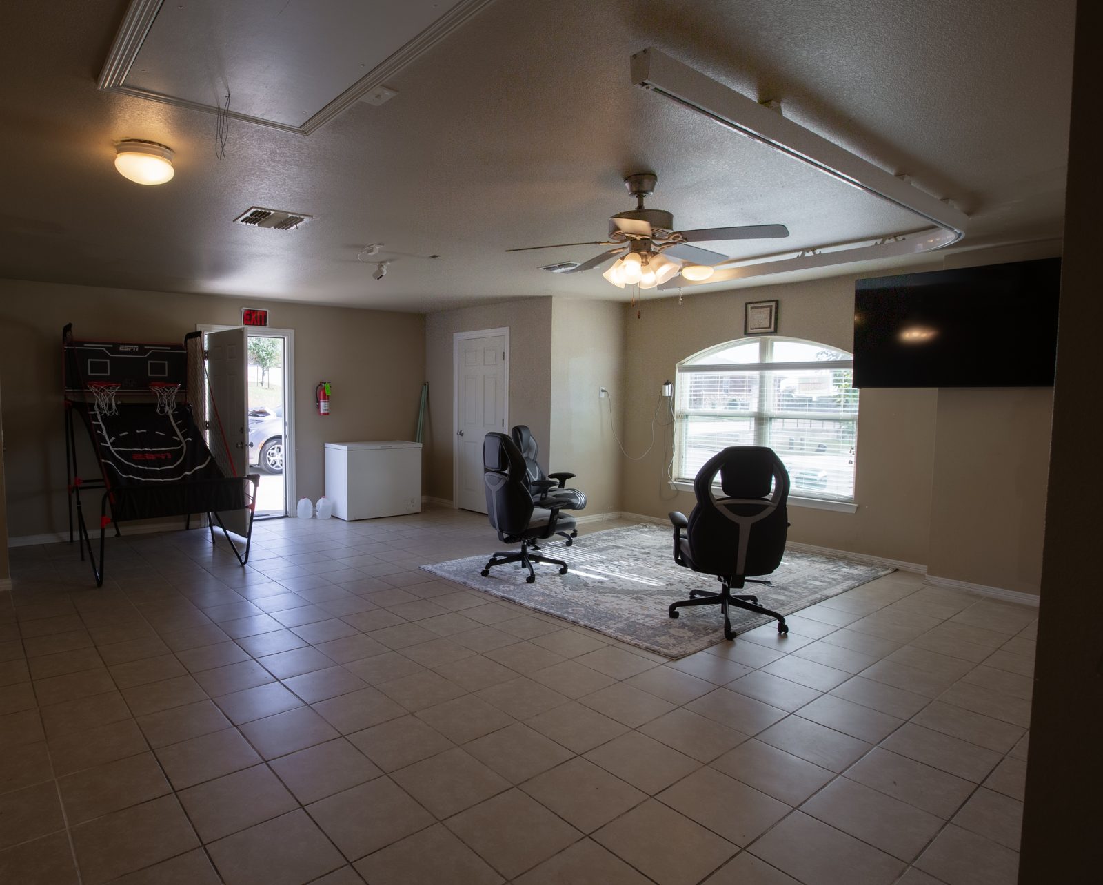 Large tiled game room with a two-hoop basketball arcade game, three gaming chairs on a rug, arched window and a big wall-mounted TV