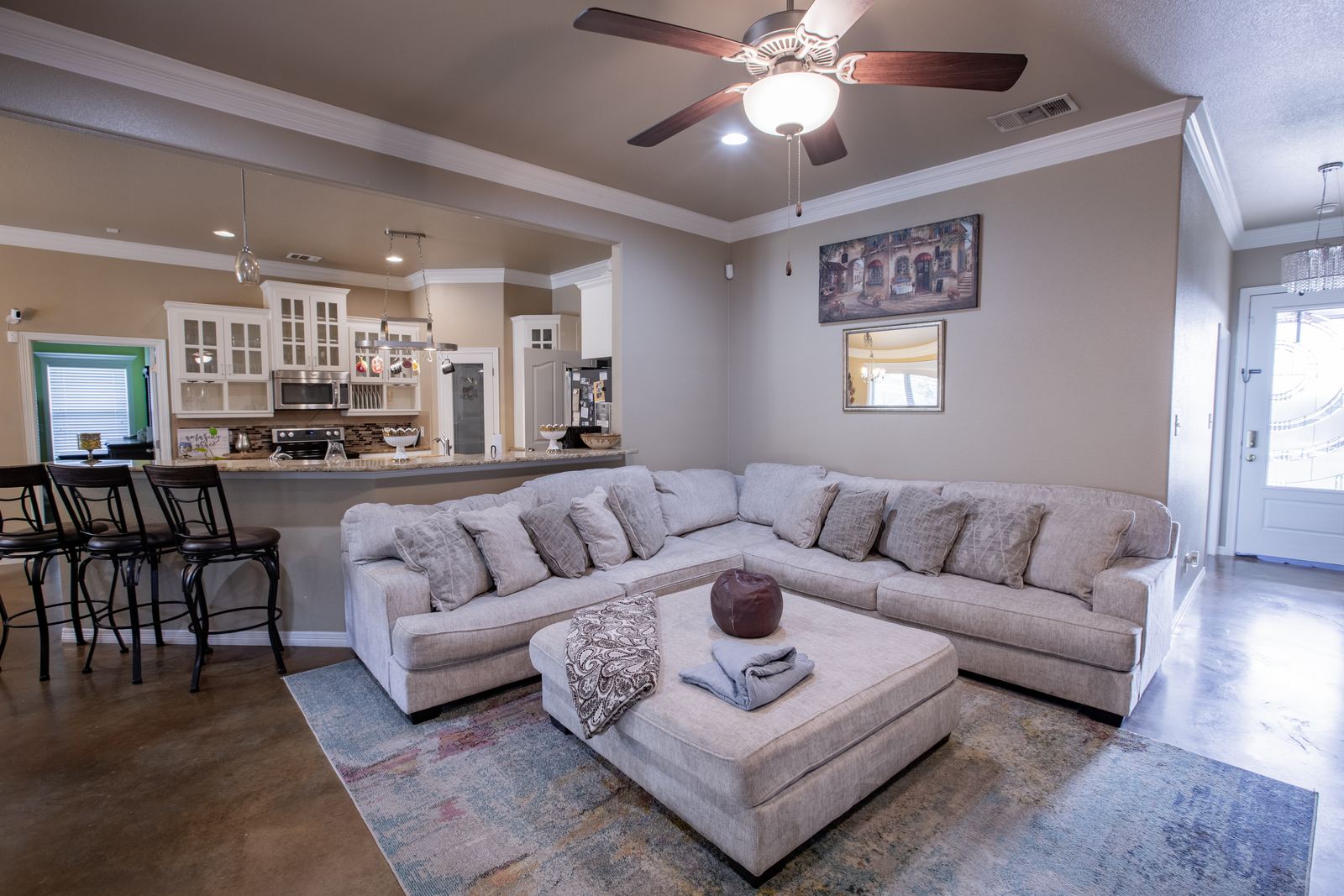 Light gray sectional and ottoman in the living room, open to a white kitchen with bar stools