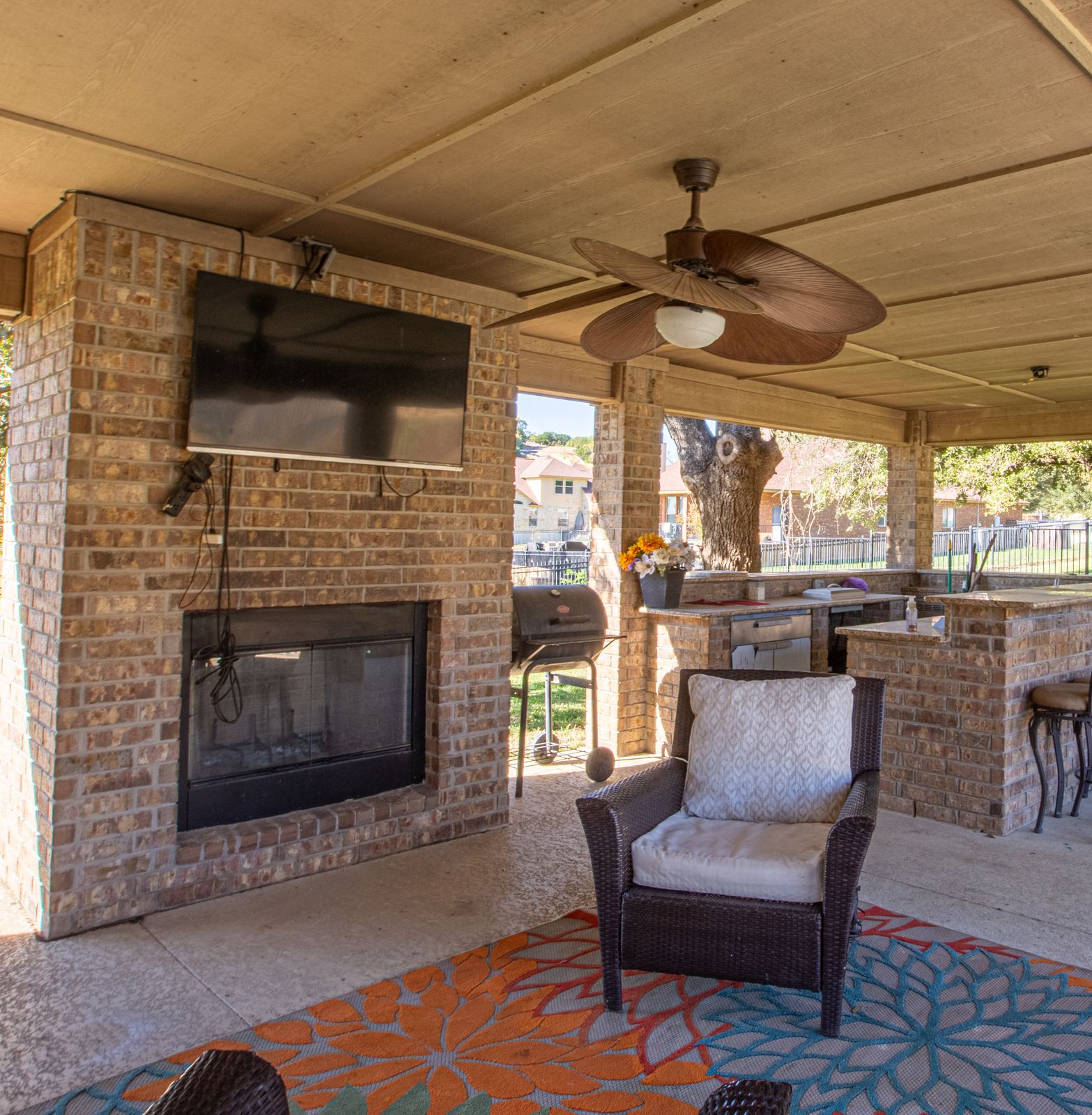 Covered brick patio with a wood-burning fireplace, wall TV, grill, outdoor kitchen bar and wicker chair on a bright rug