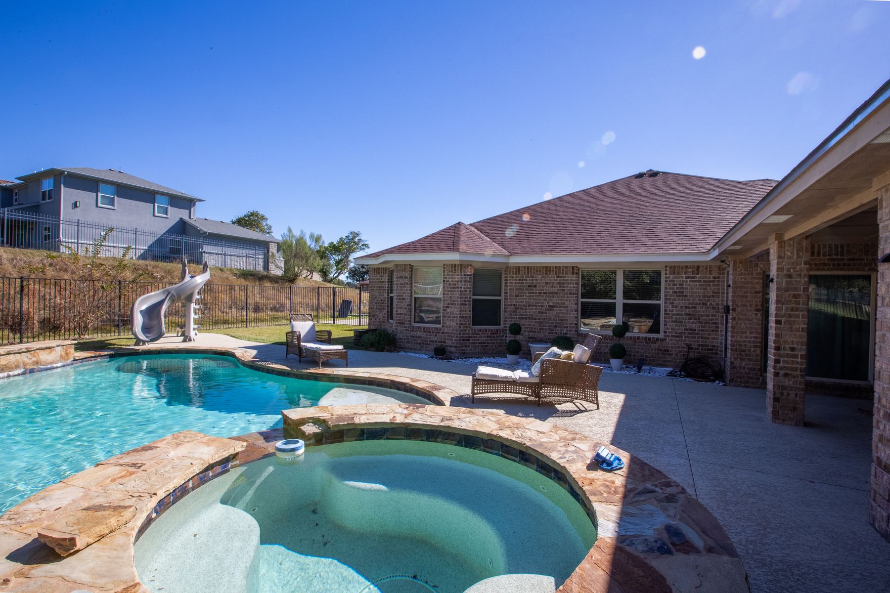 Private pool with a water slide and stone-edged jacuzzi behind the brick home under a clear blue sky
